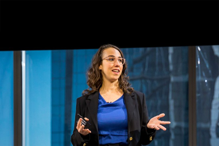 Julia Bernstein, brunette with glasses in a blue shirt and blazer speaking from a building with windows in the background